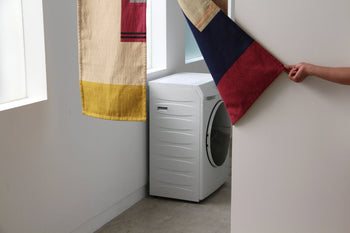A white front-loading washing machine stands near two windows, partially covered by a DETAIL Noren Curtain from DETAIL with kimono patterns as a hand pulls back one fabric panel.