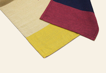 Two DETAIL Noren Curtain fabrics overlap on a light background—one with yellow and beige panels, the other with dark blue, red, and pink sections plus visible stitching. Their design recalls kimono patterns and traditional Noren curtains by DETAIL.