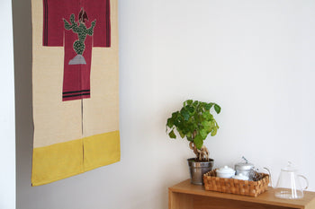 A small indoor plant in a silver pot sits on a shelf beside a basket of teapots and cups, with a glass teapot nearby. Above, a DETAIL Noren Curtain by DETAIL featuring a colorful cactus design decorates the white wall.