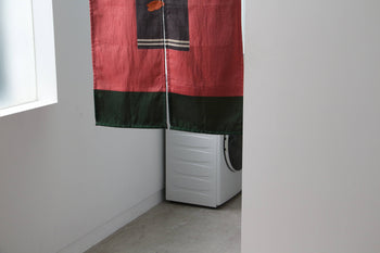 A DETAIL Noren Curtain by DETAIL, adorned with kimono patterns, hangs in a minimalistic white room with a concrete floor; a washing machine is partially visible behind it.