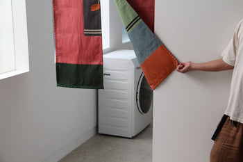 A person in a white shirt pulls aside the DETAIL Noren Curtain by DETAIL, featuring vivid kimono patterns, to reveal a white front-loading washing machine in a bright, minimal laundry room.