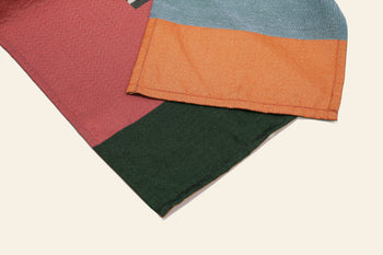 Two pieces of fabric from the DETAIL Noren Curtain by DETAIL overlap: one showcases blue and orange kimono patterns, the other has red and green sections. Both are set against a light background.
