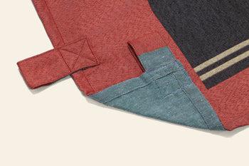 A close-up of the corner of the DETAIL Noren Curtain by DETAIL, featuring a stitched tab, folded hem with visible stitching, and a striped detail inspired by traditional kimono patterns in red and blue fabric.