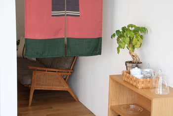 A cozy corner with a wooden armchair, partially hidden by the DETAIL Noren Curtain in pink and green, a potted plant on a shelf, and a woven basket with teapots and cups on a light wooden cabinet.