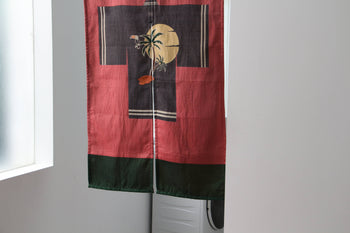 The DETAIL Noren Curtain by DETAIL features red and black panels with a yellow moon, palm trees, and a bird silhouette, its plant-inspired design softly concealing a white washing machine in the doorway.