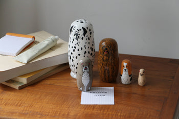 Five DETAIL Partners Nesting Dolls by DETAIL, each painted as a unique dog, are displayed on a wooden table with books, a tube, and a notepad. A small note in front reads "Welcome home.