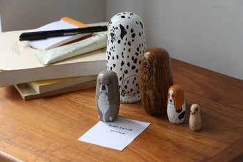 DETAIL Partners Nesting Dolls by DETAIL, featuring five hand-painted wooden dogs, are displayed on a desk next to books, pens, a notepad, and a "WELCOME HOME" card.