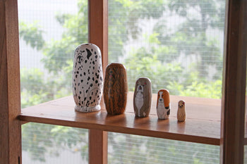The DETAIL Partners Nesting Dolls by DETAIL are five hand-painted wood dolls, each with a unique dog design, displayed in a row on a wooden shelf with a window framing green leafy trees behind them.