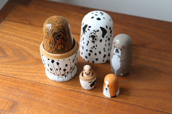 The DETAIL Partners Nesting Dolls by DETAIL are a set of five hand-painted wooden dolls, each with a unique dog design, shown separated and arranged from largest to smallest on a wooden surface.