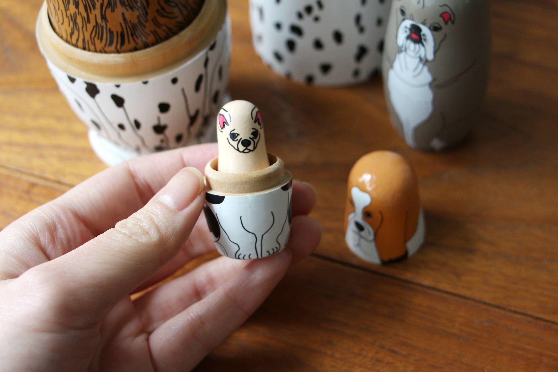 A hand holds a DETAIL Partners Nesting Doll by DETAIL, featuring a hand-painted dog design. Other dog-themed nesting dolls and pieces from the set are scattered on the wooden surface in the background.