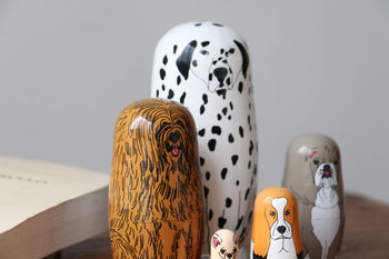 DETAIL Partners Nesting Dolls by DETAIL, each hand-painted with a unique dog design, are displayed on a table beside an open book. The largest doll features a Dalmatian.