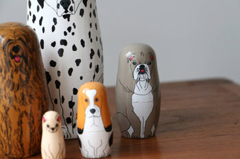 DETAIL Partners Nesting Dolls by DETAIL feature five hand-painted wood nesting dolls with charming dog designs, including a Dalmatian, Bulldog, Basset Hound, and more, displayed on a wooden surface with a gray background.
