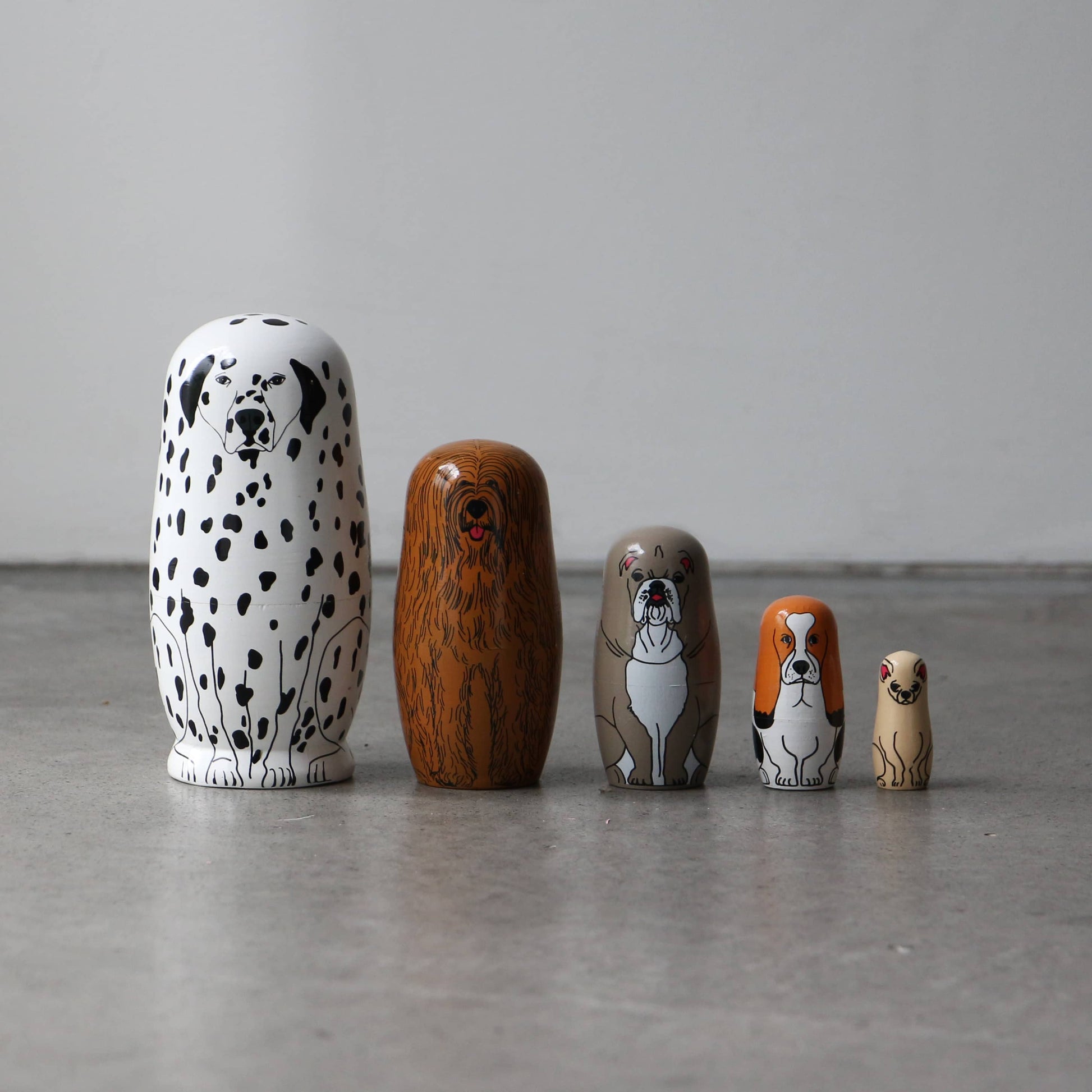 The DETAIL Partners Nesting Dolls by DETAIL feature five hand-painted wooden dolls with unique dog designs, arranged from largest to smallest on a gray floor against a plain white wall.
