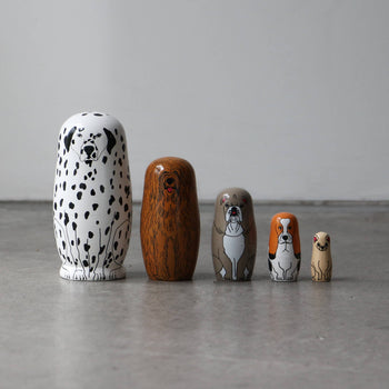 The DETAIL Partners Nesting Dolls by DETAIL feature five hand-painted wooden dolls with unique dog designs, arranged from largest to smallest on a gray floor against a plain white wall.