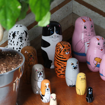 DETAIL Partners Nesting Dolls by DETAIL, featuring hand-painted cat, dog, and tiger designs, are displayed on a wooden surface beside a potted plant in a decorative room corner.