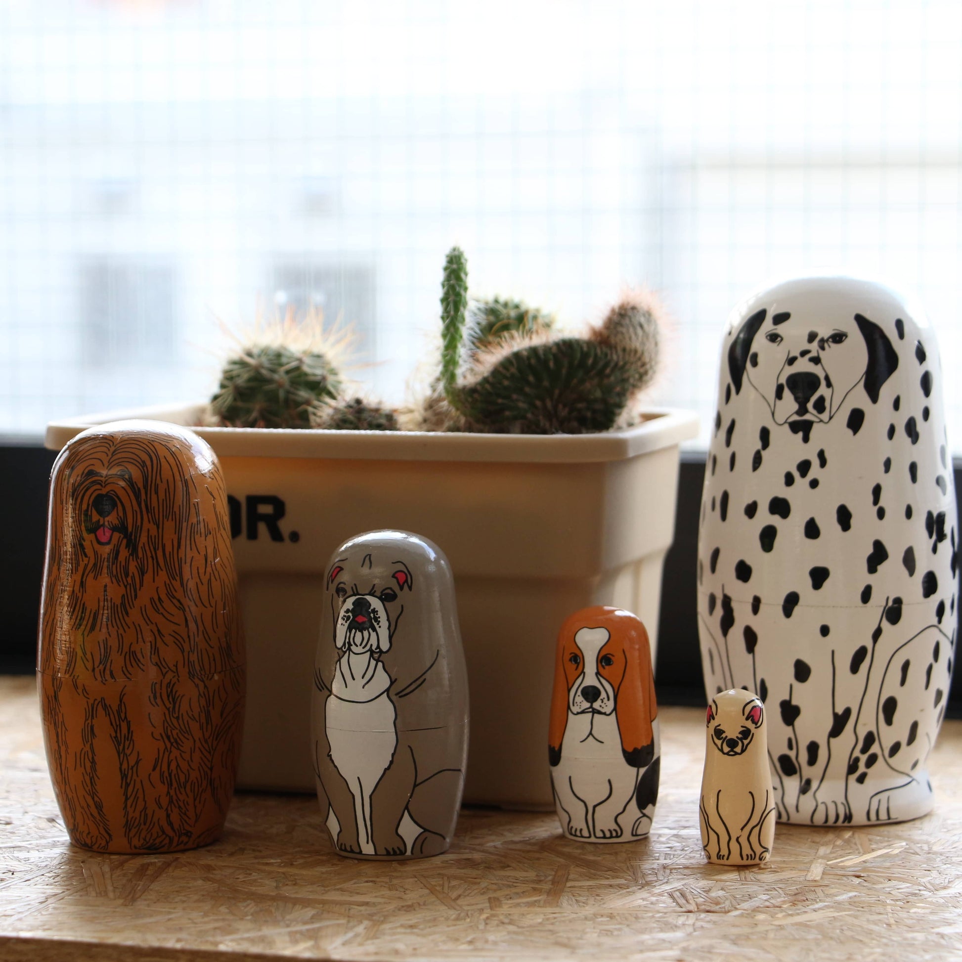 Five DETAIL Partners Nesting Dolls by DETAIL, each hand-painted with a different dog breed, are lined up in front of a beige cactus pot. A bright window with a grid pattern forms the background.