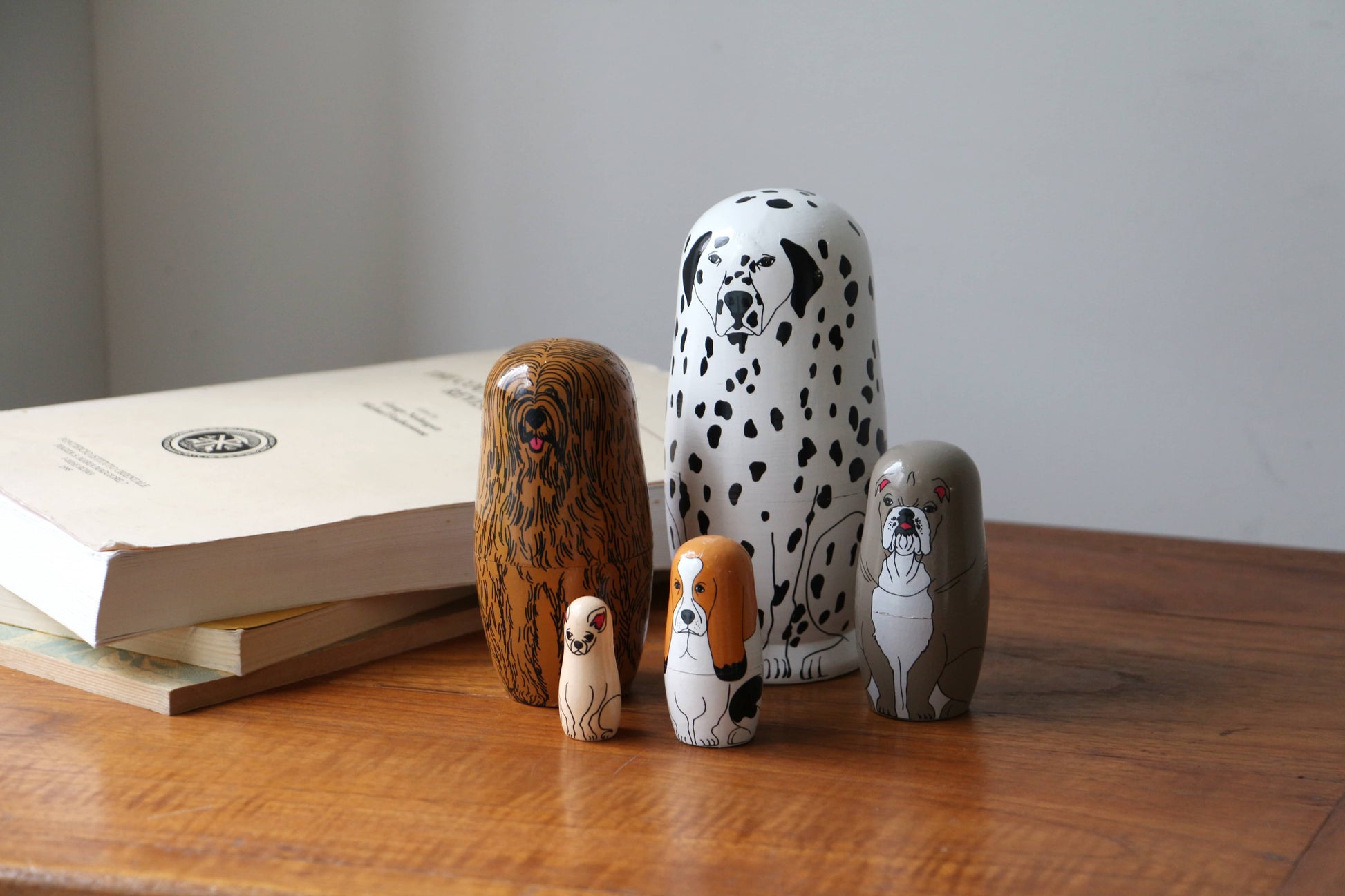 Five DETAIL Partners Nesting Dolls by DETAIL, each with a unique hand-painted dog design, are arranged on a wooden table beside stacked books against a plain, light-colored wall.