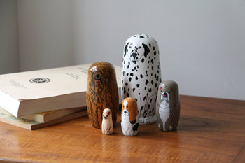 Five DETAIL Partners Nesting Dolls by DETAIL, each with a unique hand-painted dog design, are arranged on a wooden table beside stacked books against a plain, light-colored wall.