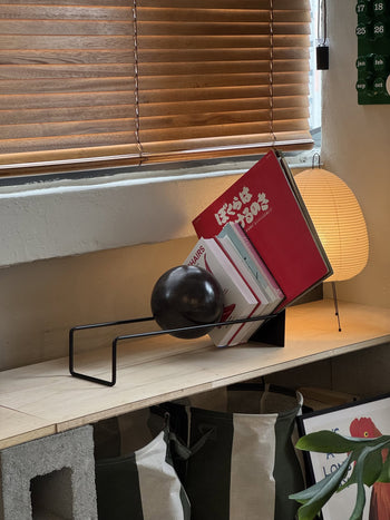 The DETAIL Slope Bookend by DETAIL, featuring a sculptural design with a spherical weight, keeps books upright on a wooden shelf beside a paper lantern under a window with partially closed blinds.