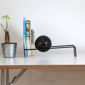 A small potted plant, several books, and the DETAIL Slope Bookend by DETAIL, featuring black metal and a dark wooden sphere, sit on a white desk against a plain white wall.
