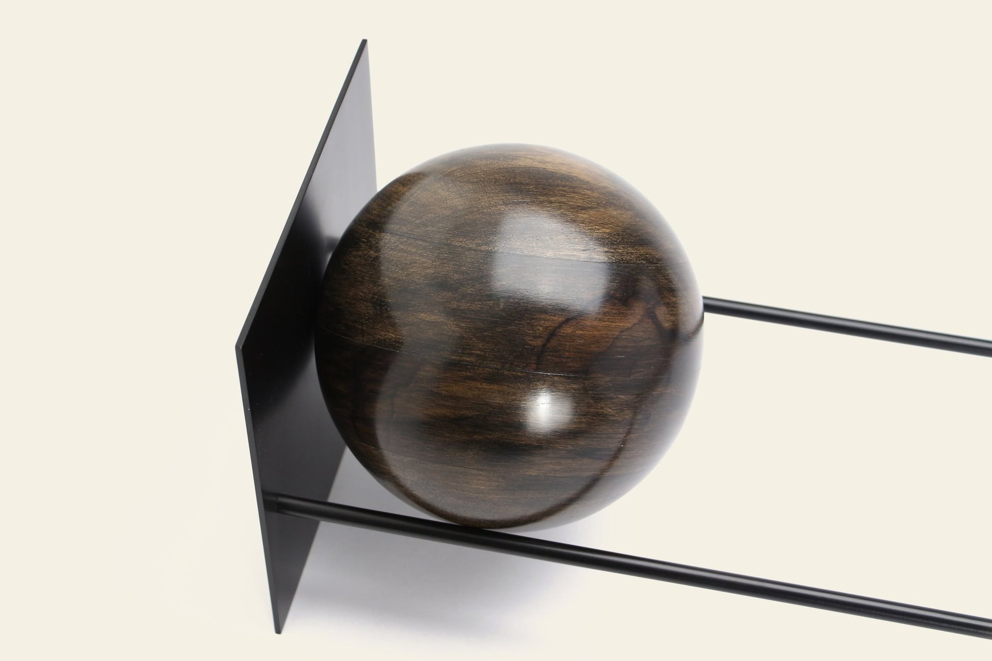 The DETAIL Slope Bookend by DETAIL features a polished dark wooden sphere set between two parallel black metal rods and a vertical black metal plate, forming a sculptural accent against a light backdrop.
