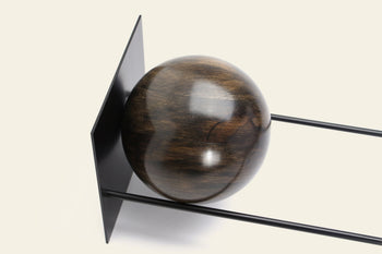 The DETAIL Slope Bookend by DETAIL features a polished dark wooden sphere set between two parallel black metal rods and a vertical black metal plate, forming a sculptural accent against a light backdrop.