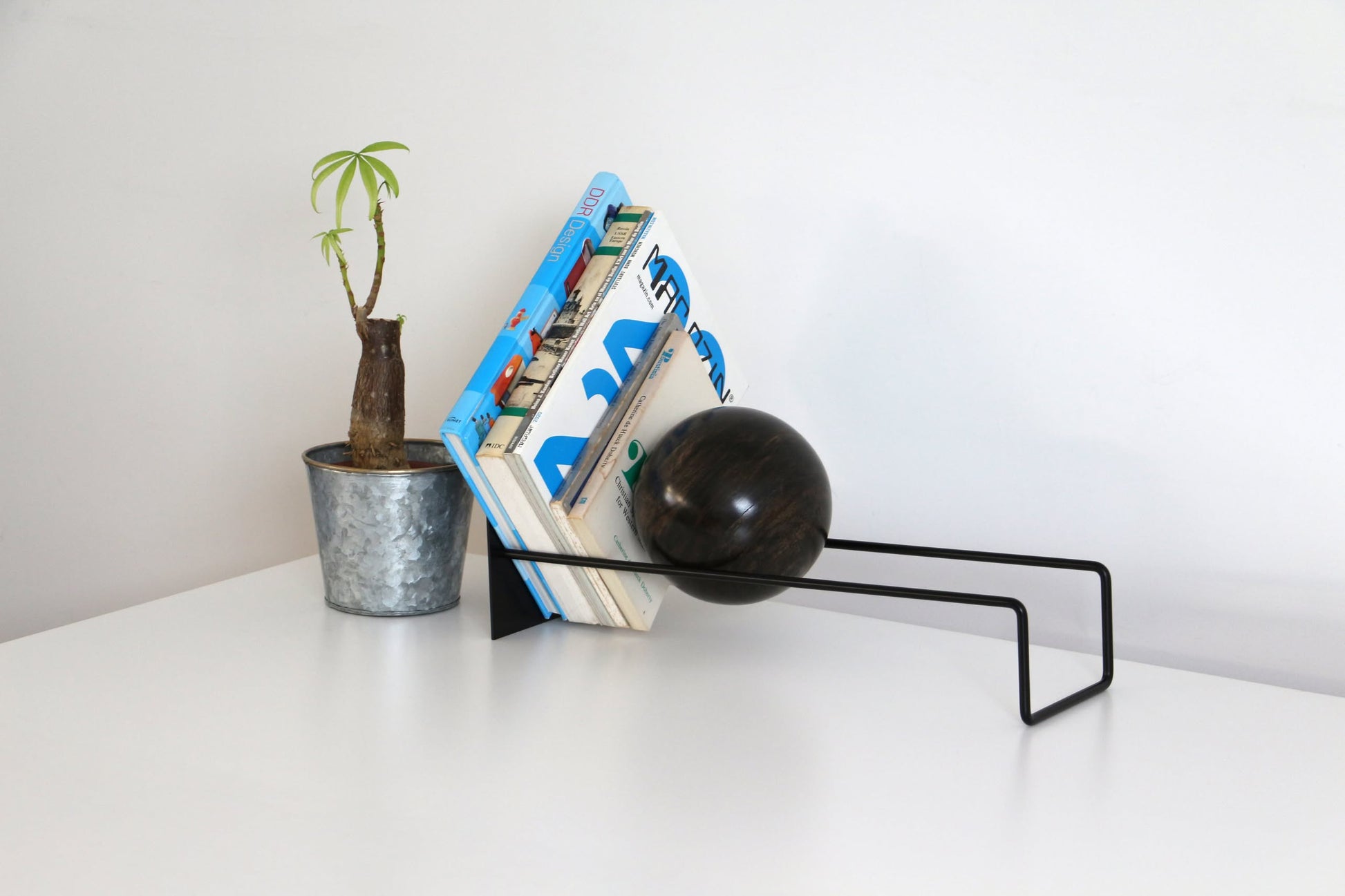 A DETAIL Slope Bookend by DETAIL with a dark sphere and a small potted plant hold up books on a white desk against a plain, light wall.