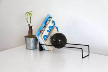 A DETAIL Slope Bookend by DETAIL with a dark sphere and a small potted plant hold up books on a white desk against a plain, light wall.