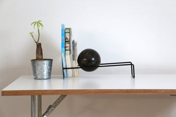 A small potted plant, upright books, and the DETAIL Slope Bookend by DETAIL are arranged on a minimalist white desk with a wooden edge against a plain white wall.