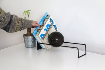 A person arranges books in the DETAIL Slope Bookend by DETAIL, featuring a black metal frame and wooden sphere, beside a potted plant on a white surface.