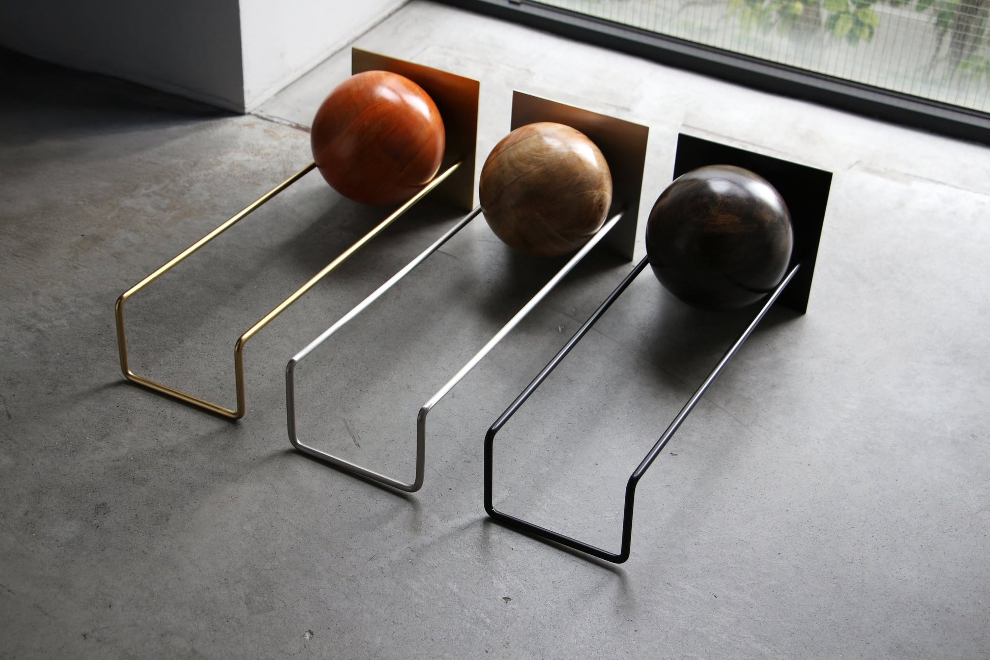 Three DETAIL Slope Bookends by DETAIL sit by a window on a concrete floor, each with a metal frame and wooden sphere: gold with light brown, silver with tan, and black with dark brown.
