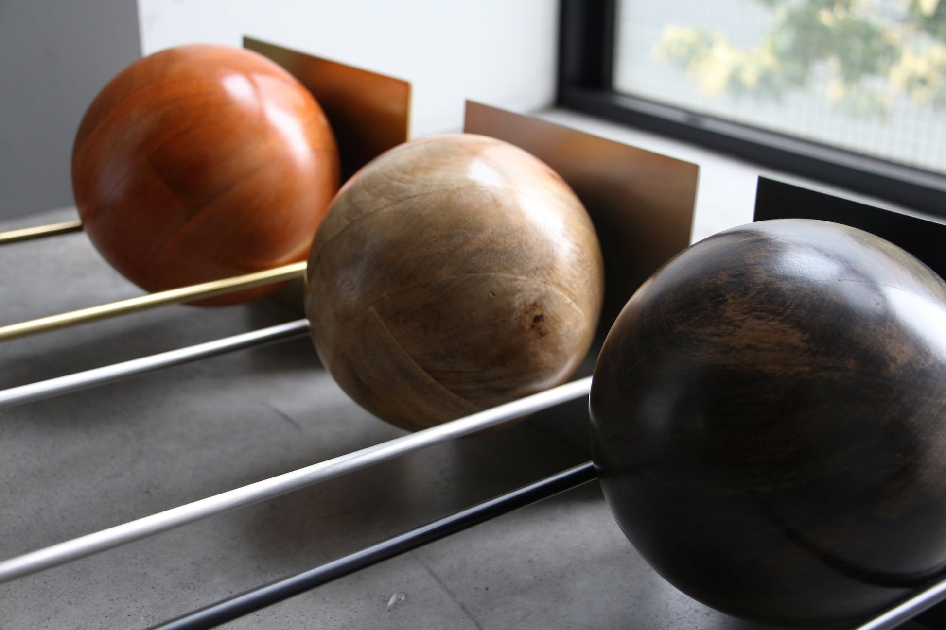 The DETAIL Slope Bookend by DETAIL features three large wooden spheres in brown, tan, and dark brown, mounted on metal rods with rectangular backings for a sculptural display. Shown on a concrete floor near a window.