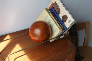 The DETAIL Slope Bookend by DETAIL, a sculptural wooden ball, holds books in a metal stand on wood, sunlight casting shadows. In the background, a wooden chair with dark fabric draped over it is partly visible.