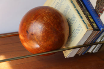 The DETAIL Slope Bookend by DETAIL, a polished wooden sphere with sleek metal rods, supports upright books on a shelf. Sunlight casts soft shadows across this minimalist and serene scene.