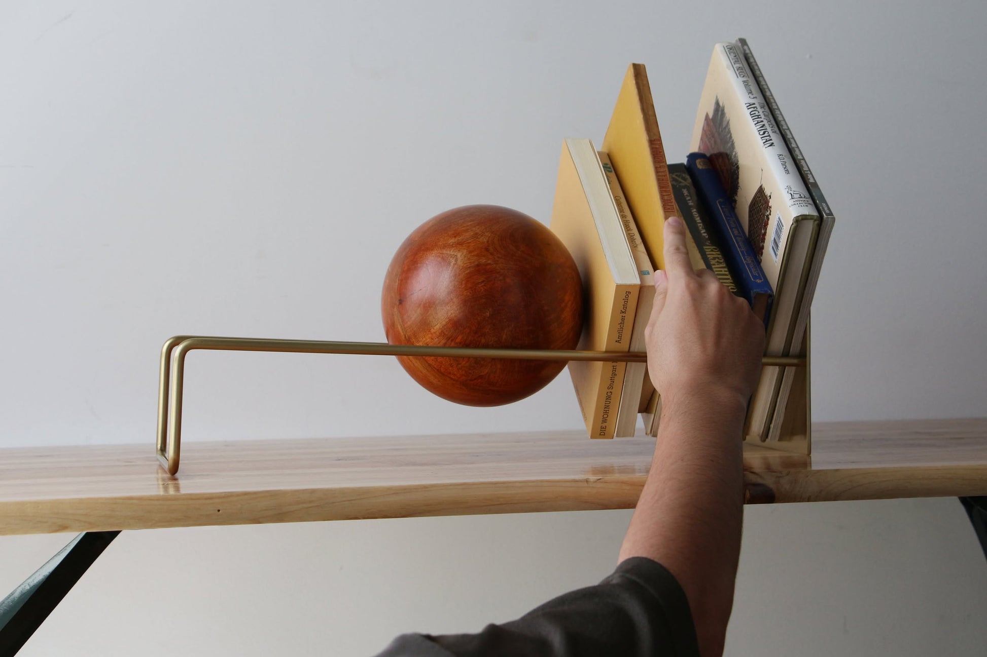 A hand reaches for a book from a stylish wooden and metal bookshelf, featuring the DETAIL Slope Bookend by DETAIL—a large orange sculptural piece—used as a bookend, with several neatly arranged books on the shelf.