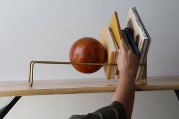 A hand reaches for a book from a stylish wooden and metal bookshelf, featuring the DETAIL Slope Bookend by DETAIL—a large orange sculptural piece—used as a bookend, with several neatly arranged books on the shelf.