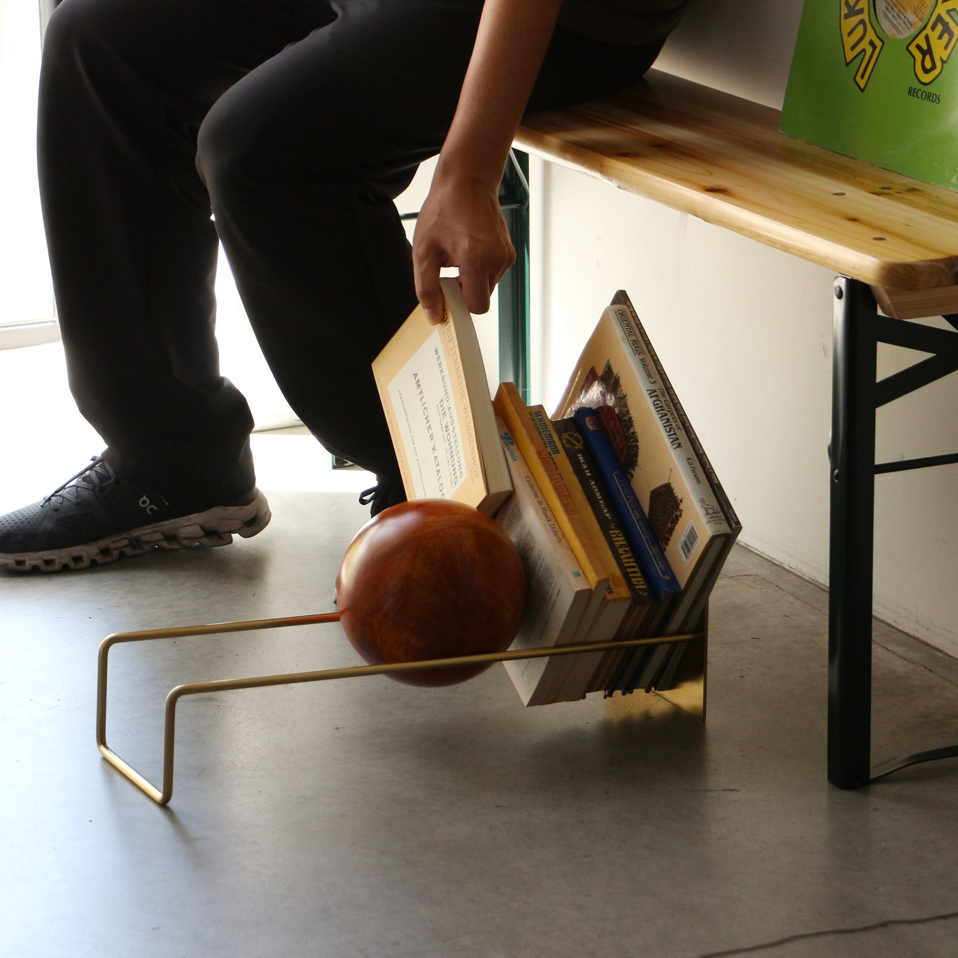 A person sitting on a wooden bench picks up a book from the DETAIL Slope Bookend by DETAIL, a sculptural metal book holder on the floor featuring several books and a wooden ball accent in its minimalist design.