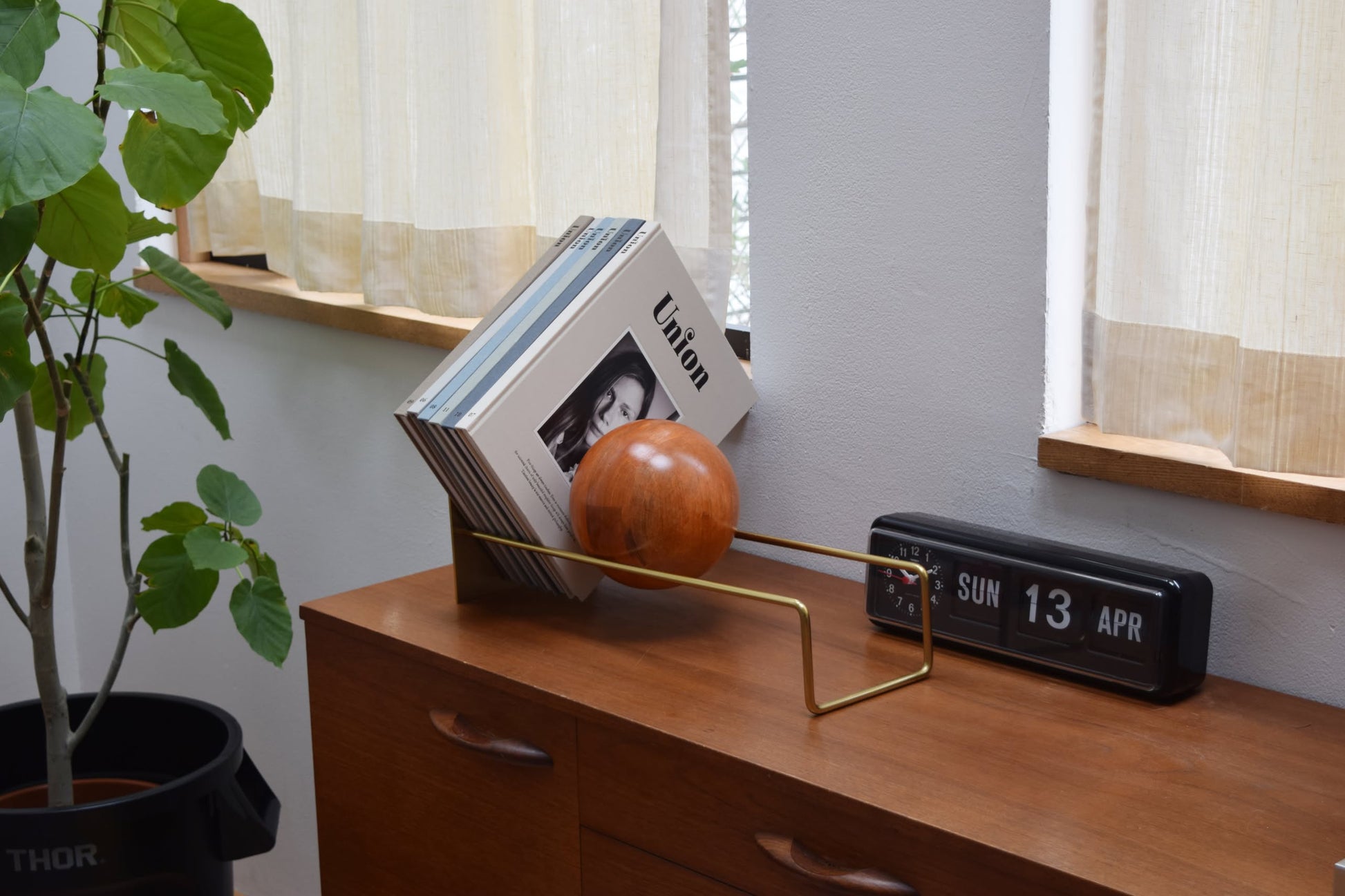 A wooden sideboard with a modern vinyl record holder, several records, the DETAIL Slope Bookend by DETAIL, a flip clock showing SUN 13 APR, a leafy potted plant, and sunlight streaming through curtains.