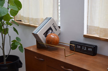A wooden sideboard with a modern vinyl record holder, several records, the DETAIL Slope Bookend by DETAIL, a flip clock showing SUN 13 APR, a leafy potted plant, and sunlight streaming through curtains.