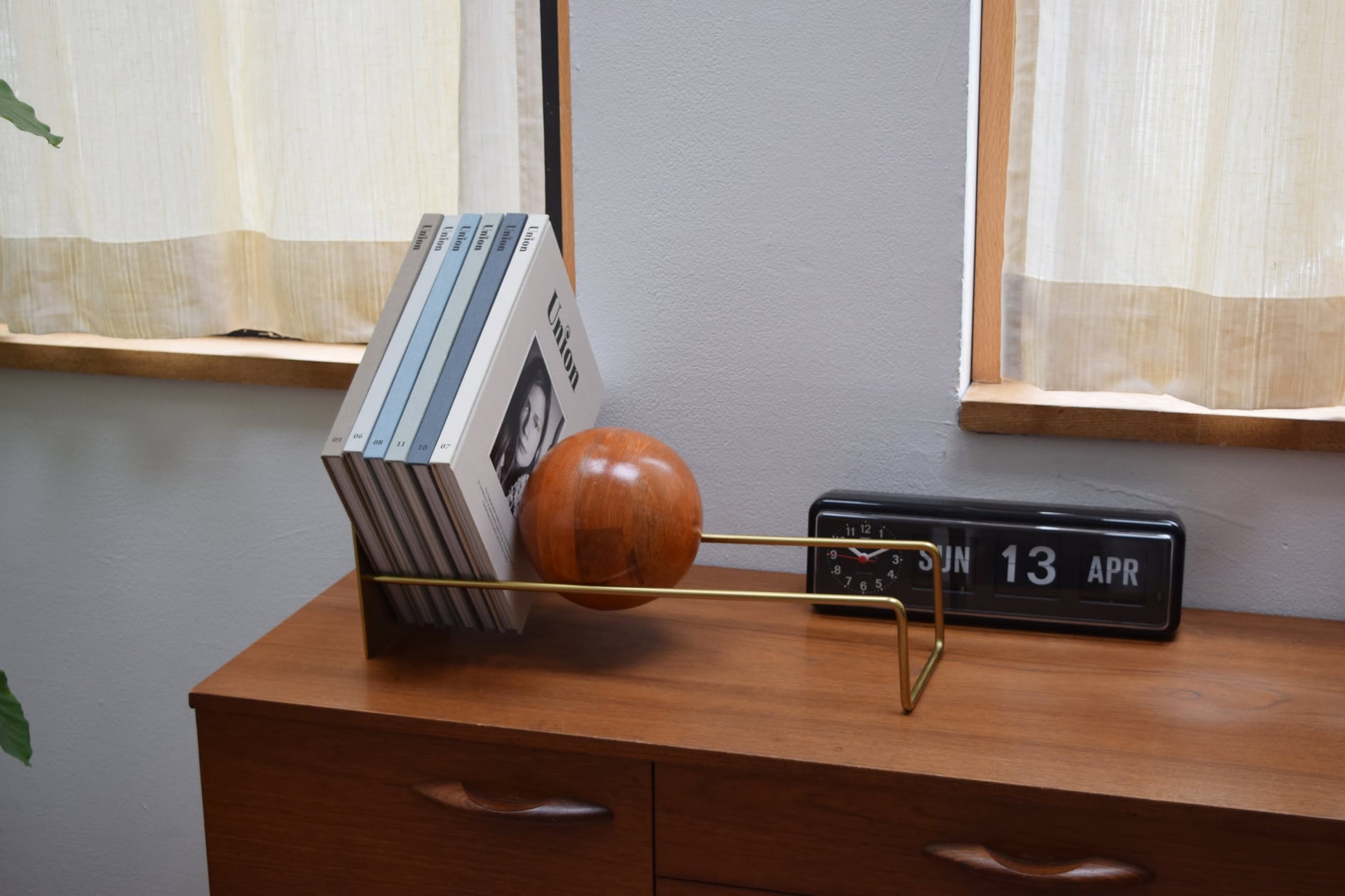 A modern wooden sideboard holds a vinyl record holder with five records and the DETAIL Slope Bookend by DETAIL, shaped like a wooden sphere. Next to it, a black flip clock displays JUN 13 APR as light streams through nearby windows.