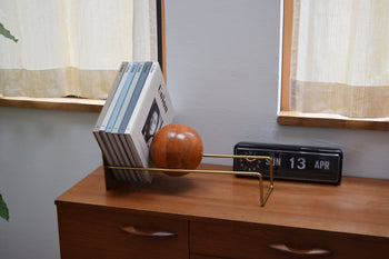 A modern wooden sideboard holds a vinyl record holder with five records and the DETAIL Slope Bookend by DETAIL, shaped like a wooden sphere. Next to it, a black flip clock displays JUN 13 APR as light streams through nearby windows.
