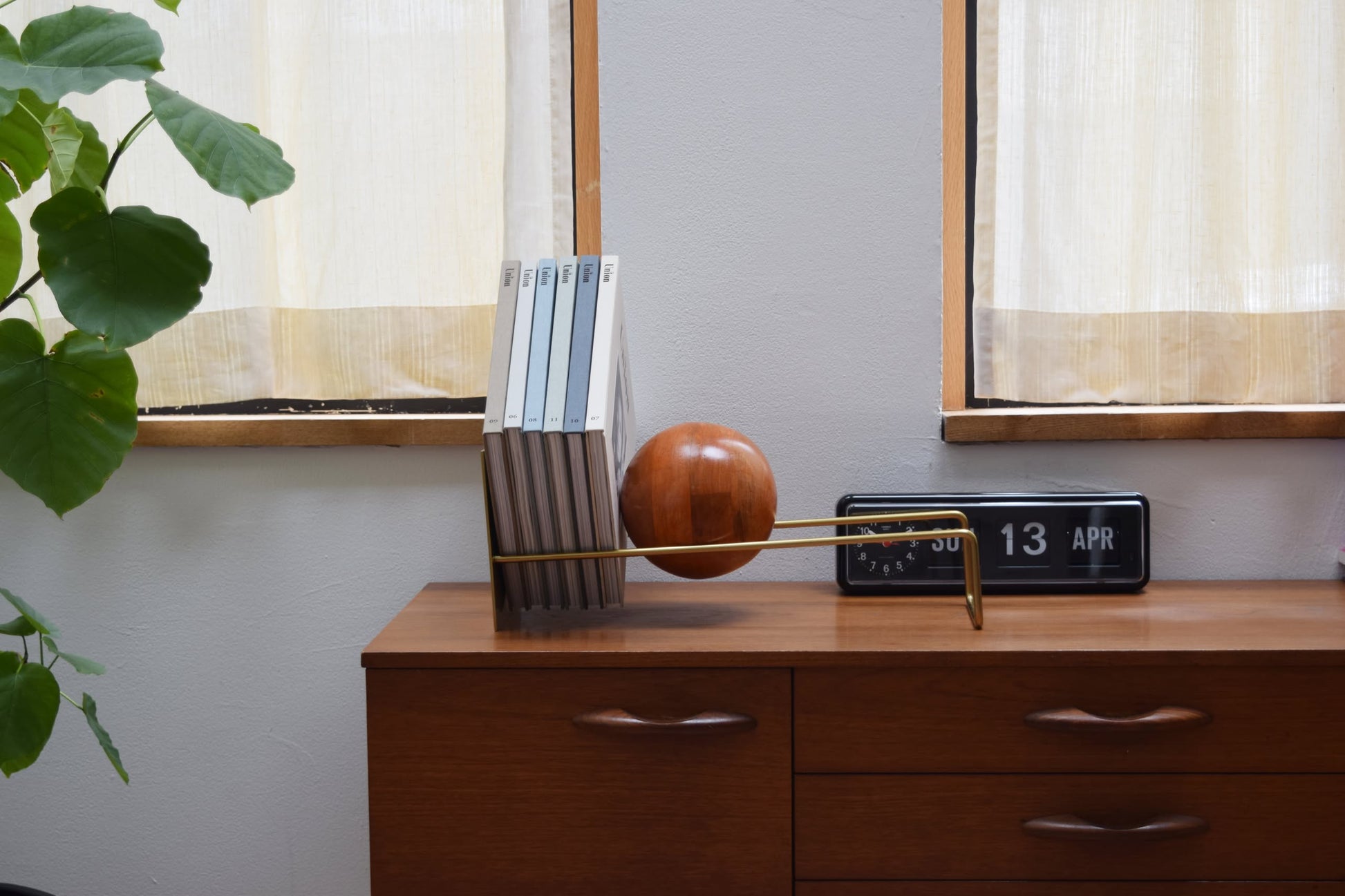 A DETAIL Slope Bookend by DETAIL with a gold rod and wooden sphere supports books on a wooden sideboard beside a black flip clock showing 13 APR, placed in front of curtained windows and a leafy green plant.