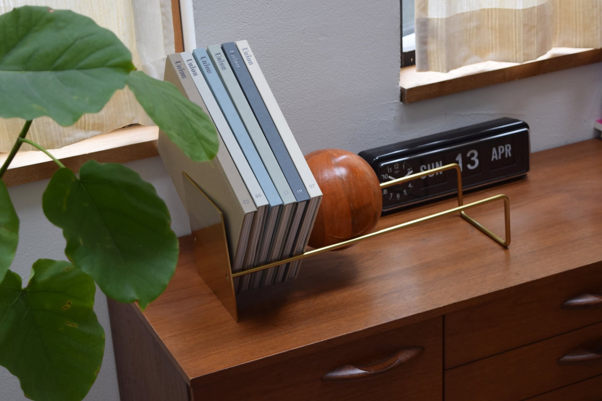 A wooden sideboard features the DETAIL Slope Bookend by DETAIL holding books, a brass magazine holder, a round wooden decor piece, a flip clock displaying SUN 13 APR, and a large green plant leaf on the left. Sunlight shines in from nearby windows.