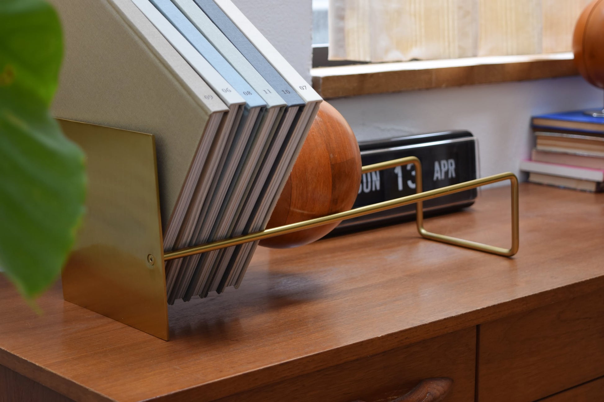 The DETAIL Slope Bookend by DETAIL, in brass, keeps books upright on a wooden desk. In the foreground is a leaf; nearby, a retro flip clock, round wood object, and stacked books sit by the window.
