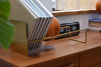 The DETAIL Slope Bookend by DETAIL, in brass, keeps books upright on a wooden desk. In the foreground is a leaf; nearby, a retro flip clock, round wood object, and stacked books sit by the window.
