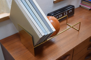 The DETAIL Slope Bookend by DETAIL in gold metal holds six books on a wooden desk, beside a brown wooden sphere and a black retro flip clock showing SUN 13 APR.