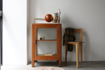 A wooden cabinet with glass doors displays a book and two decor pieces supported by the DETAIL Slope Bookend. A book holder and globe sit on top. Nearby, a wooden chair draped with a plaid blanket rests against a white wall.