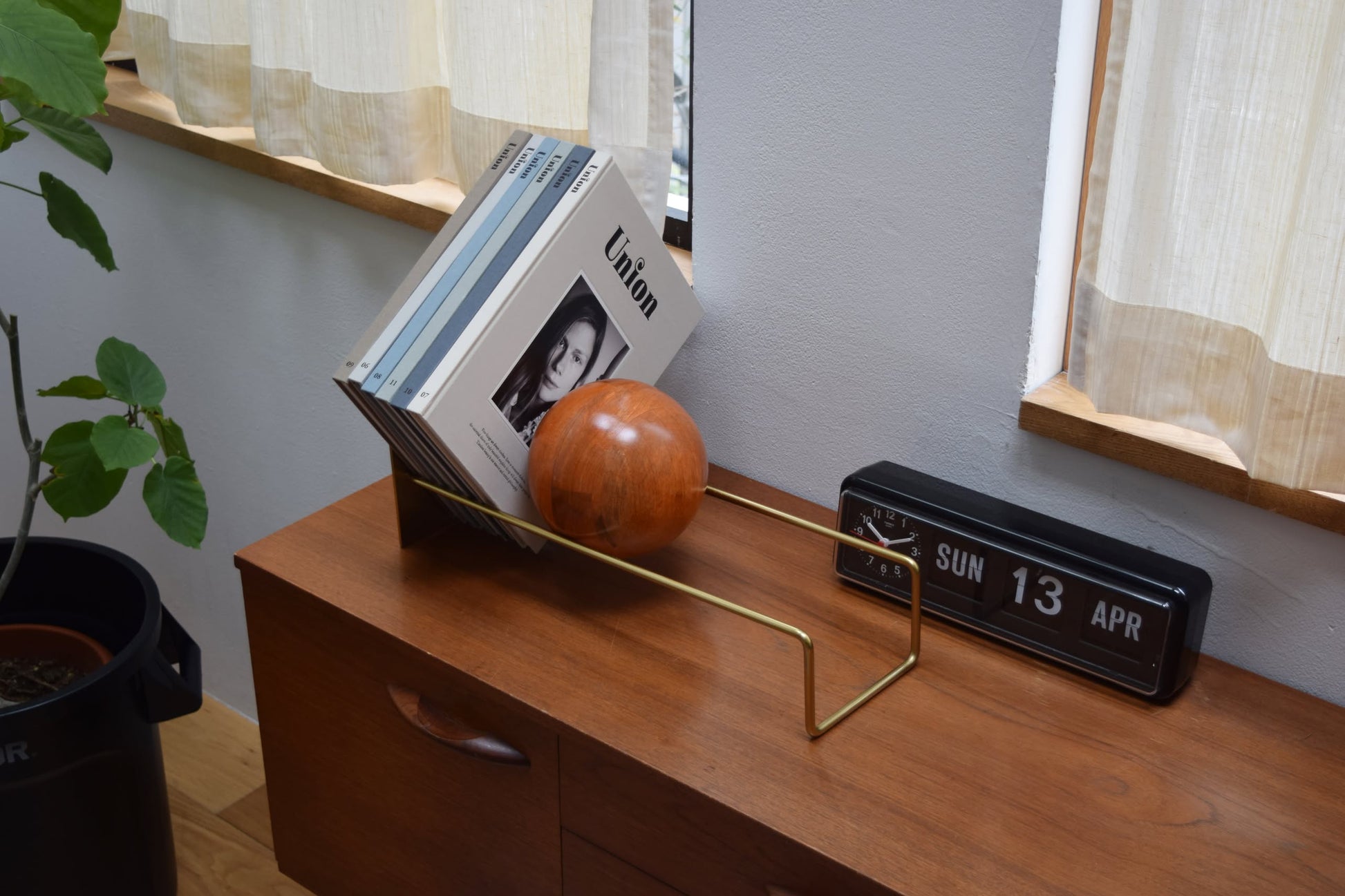 A wooden sideboard displays the DETAIL Slope Bookend by DETAIL with books and a wooden sphere, beside a retro flip clock set to Sunday, April 13. Sunlight pours through cream curtains from nearby windows.