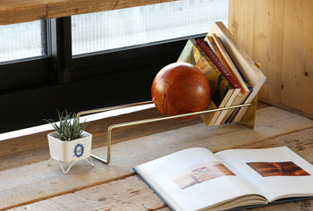 The DETAIL Slope Bookend by DETAIL holds books and a round ornament on a wooden surface; nearby are a small potted succulent and an open photo book, all arranged next to a window.