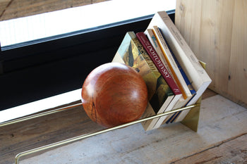 The DETAIL Slope Bookend by DETAIL holds books upright on a modern metal and wood shelf beside a window, with natural light streaming in. Its sculptural, wooden sphere design adds style to your space.